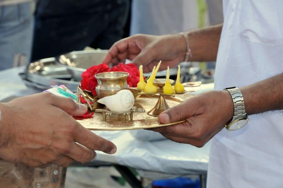 Hands holding a brass aarti plate with flowers, a conch, and oil lamps